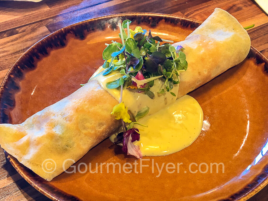 A rolled-up crepe is garnished with sprouts and topped with cheese sauce.