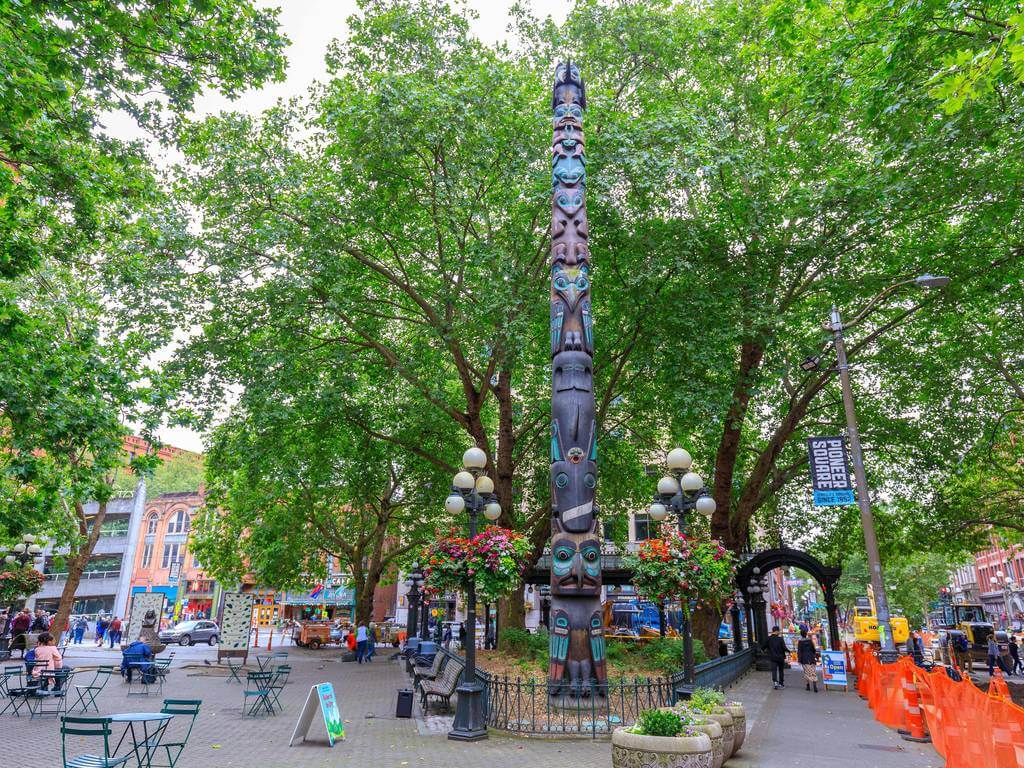 An area with a Totem Pole erected under the shades of green leaves has colorful buildings in its background.