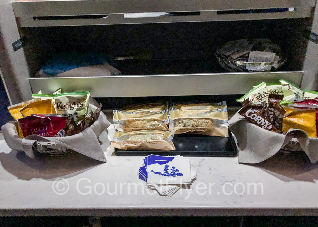 Baguette sandwiches and bags of chips and snacks on a counter in the galley.