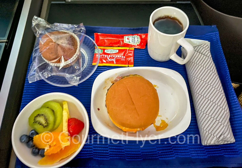 A pre-arrival tray with a burger, fruits, pastries, and a cup of black coffee.