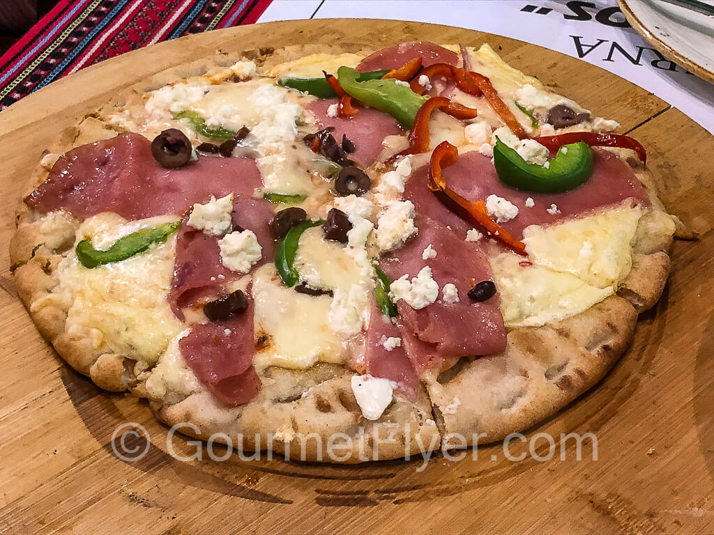 A pizza topped with smoked pork, olives, feta, tomatoes, and green peppers.