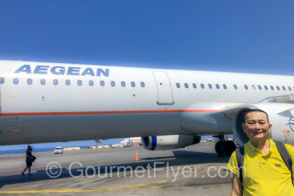 Aegean Airlines Economy Review features the Gourmet Flyer posing in front of an Aegean plane on the tarmac.