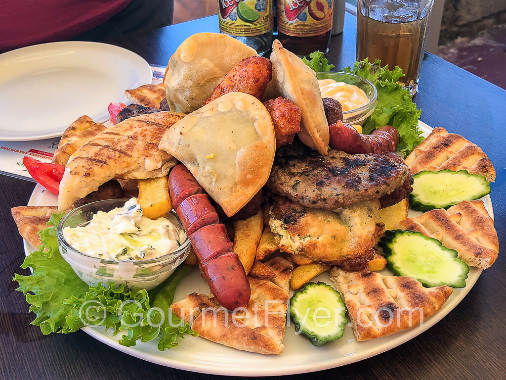 A mixed plate with a large variety of foods served with dips, sauces, and slices of cucumbers.