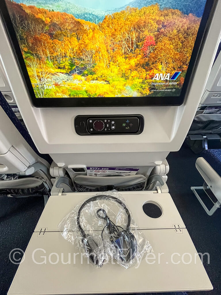Seatback entertainment screen with headphones in plastic wrap on the opened tray table.