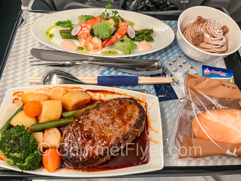 A dinner tray with a plate of hamburger steak with vegetables and potatoes, and accompanied by shrimp appetizer and dessert.