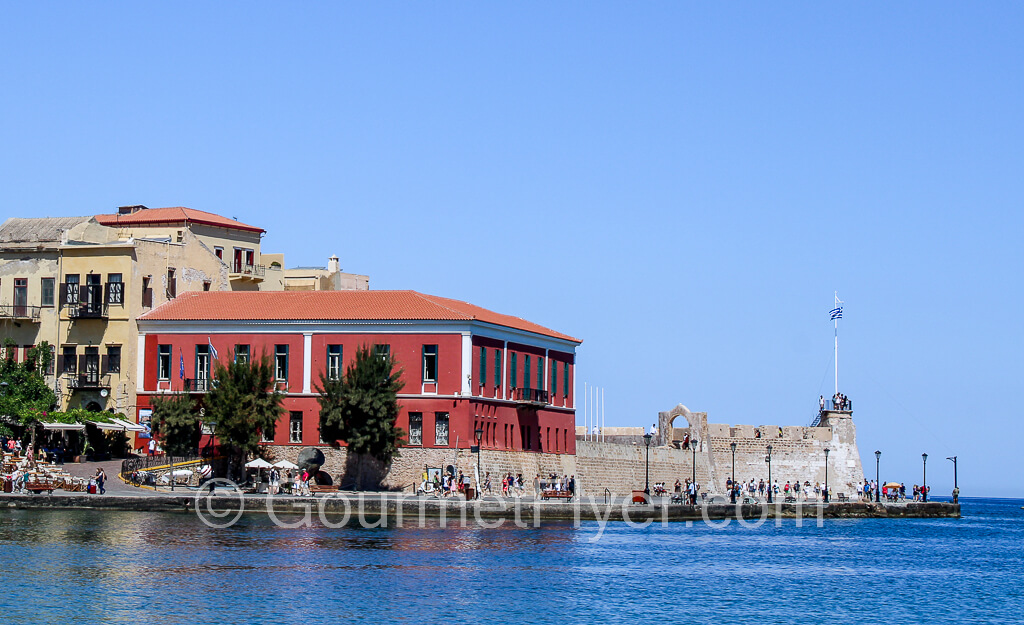 The Firka Fortress sits at the edge to the entrance to the harbor, with a brick red building next to it.