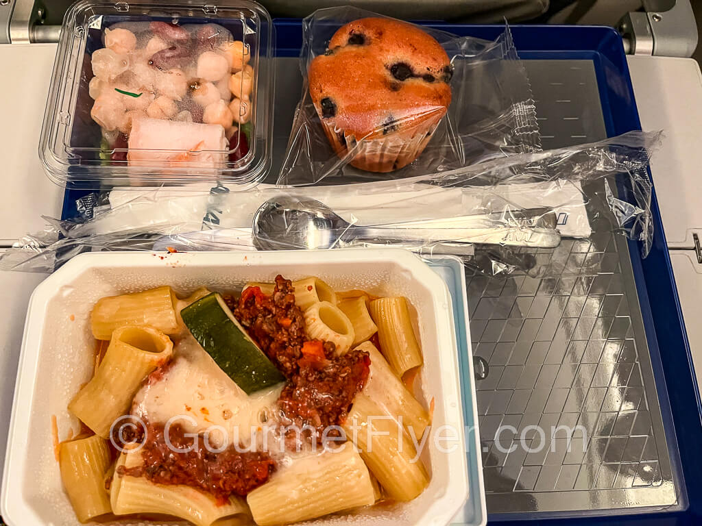 A plate of rigatoni with Bolognese meat sauce is served on a tray with a bean salad and a muffin.