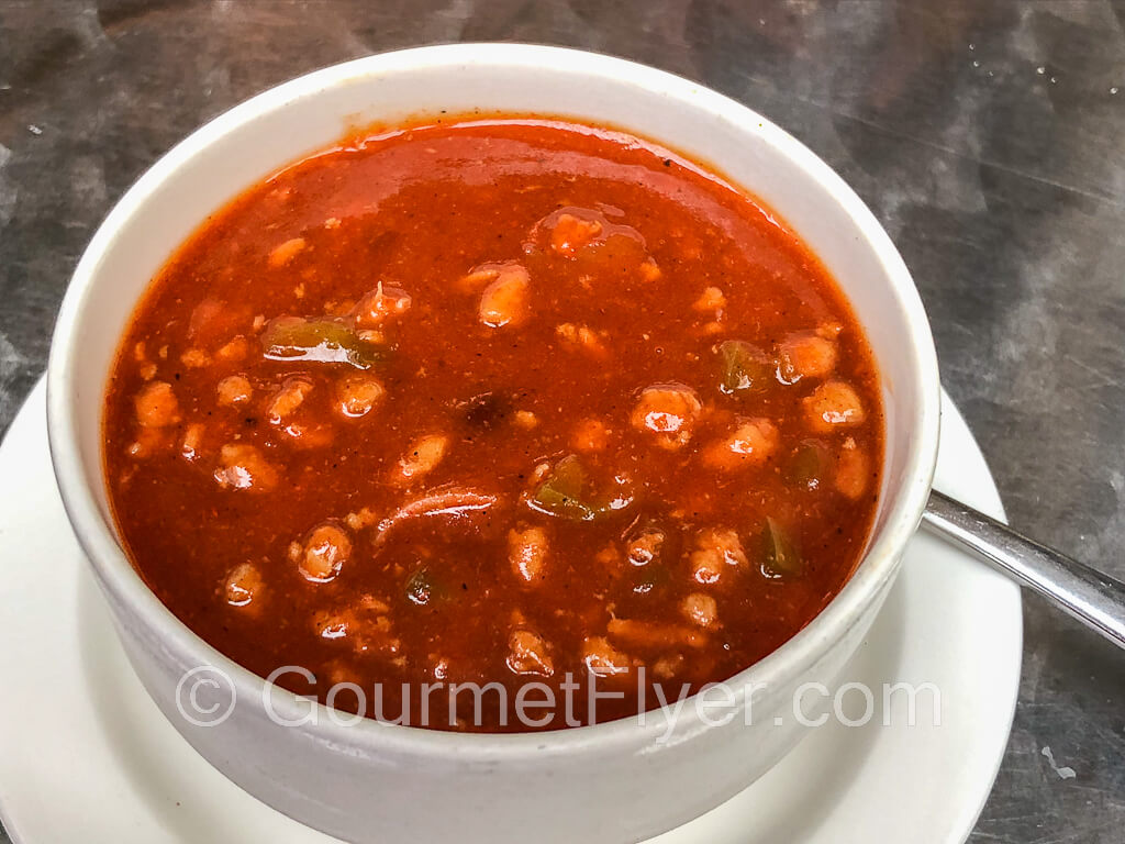 A bowl of chili on a plate showing plenty of ground beef and beans in it.