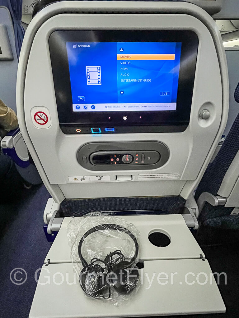 A seatback with the entertainment screen and remote control on the panel. A pair of headphones lie on top of the tray table.