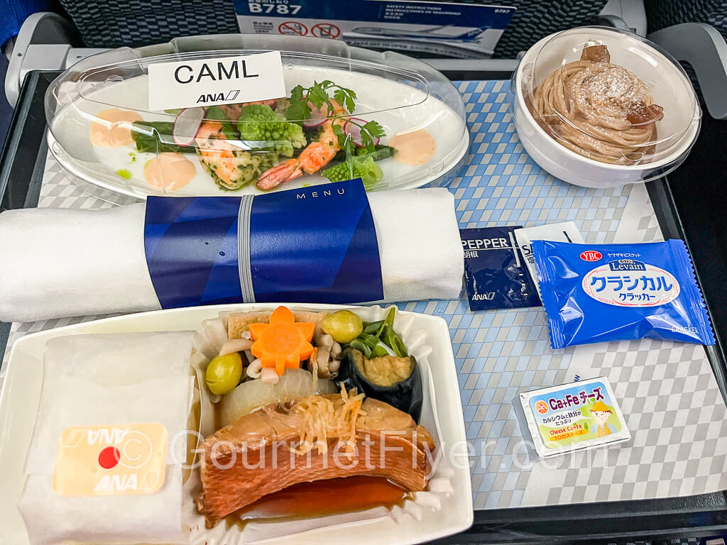 Dinner tray includes the fish entree with side dishes, shrimp appetizers, and dessert. A roll up of silverware lies on the center of the tray.