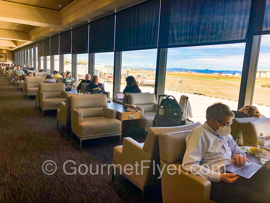 Lounge chairs with passengers line the floor to ceiling windows with tarmac view.