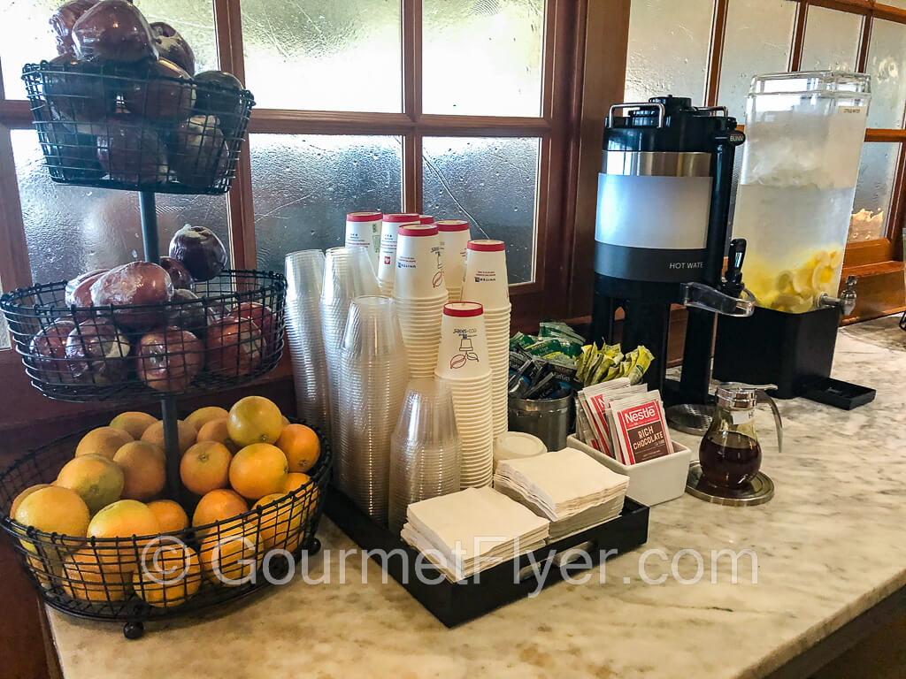 Drink station with fruits on the left, followed by a stack of cups, hot water, and iced water at the far right.