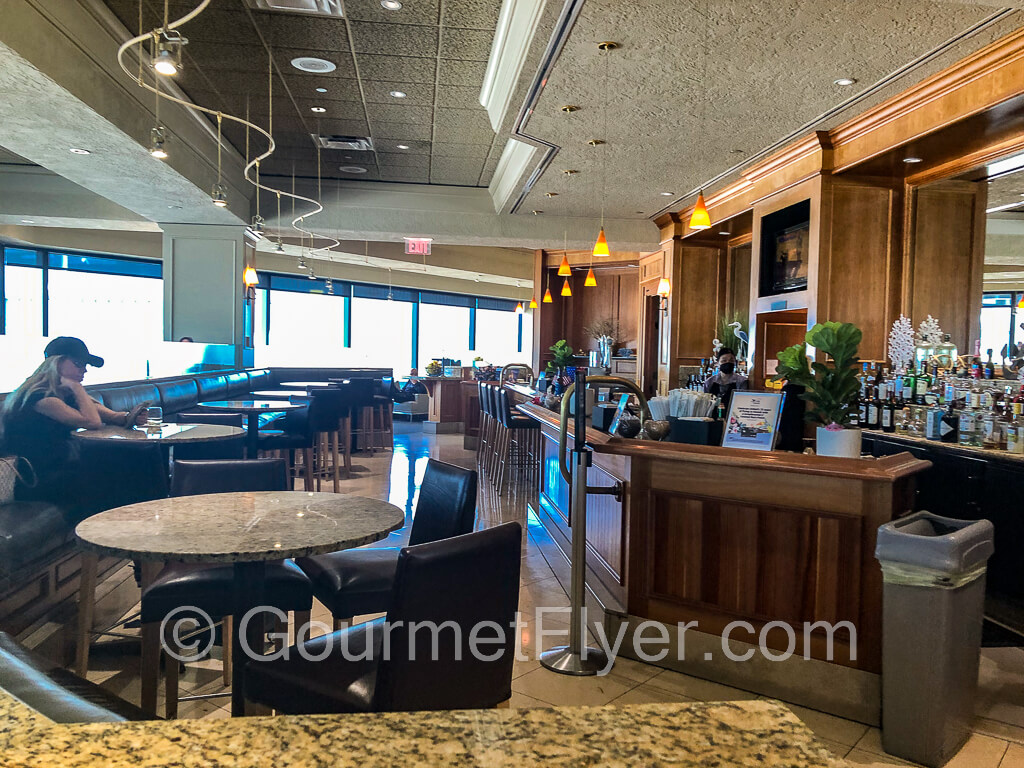 A lone customer with long blonde hair sits at a high-top table in the bar area with a full bar and many bar tables,