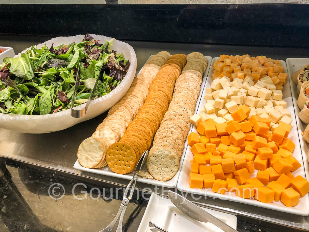 A salad bowl filled with lettuce is accompanied by 3 rows of crackers and a plate of cubes cheese.