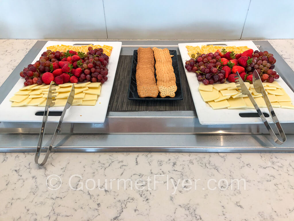 Two trays of cheese slices garnished with fresh strawberries and grapes with a plate of butterfly crackers positioned between them.