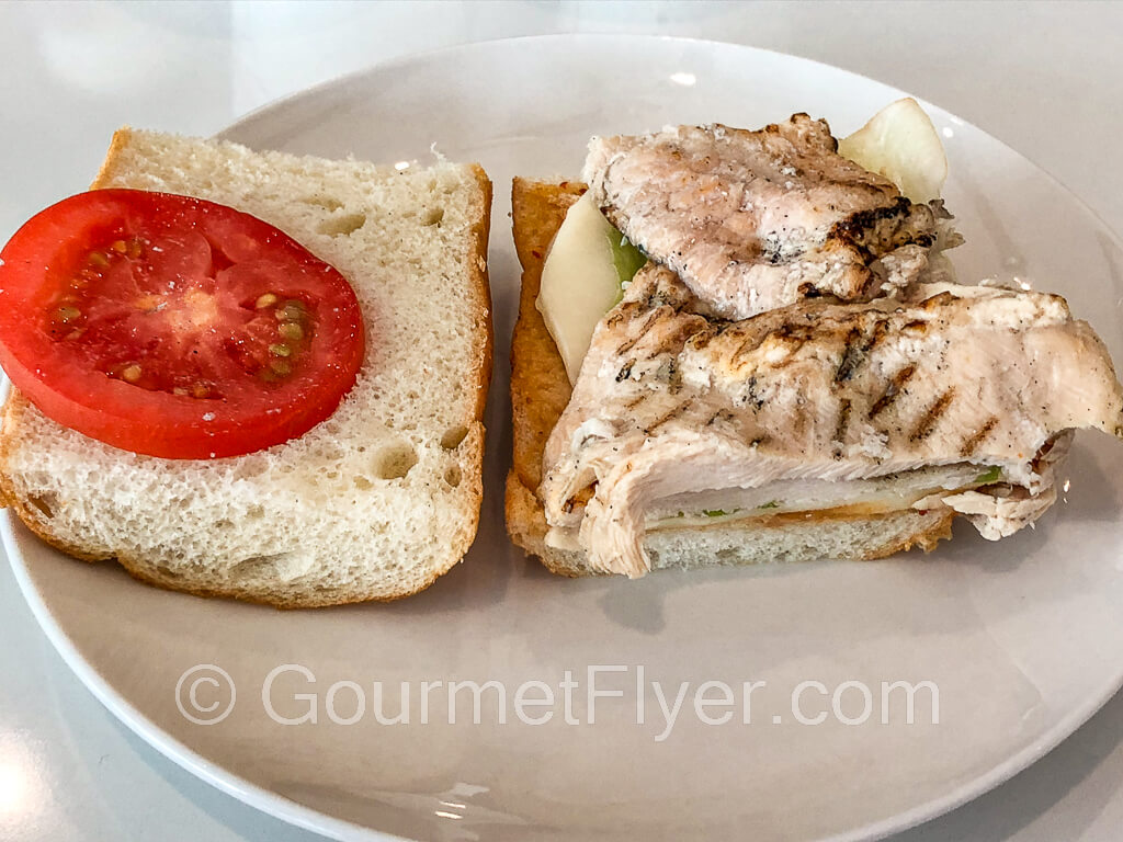 A baguette sandwich opened up to expose the grilled chicken breast sitting on cheese on one side and a slice of tomato on the other side.