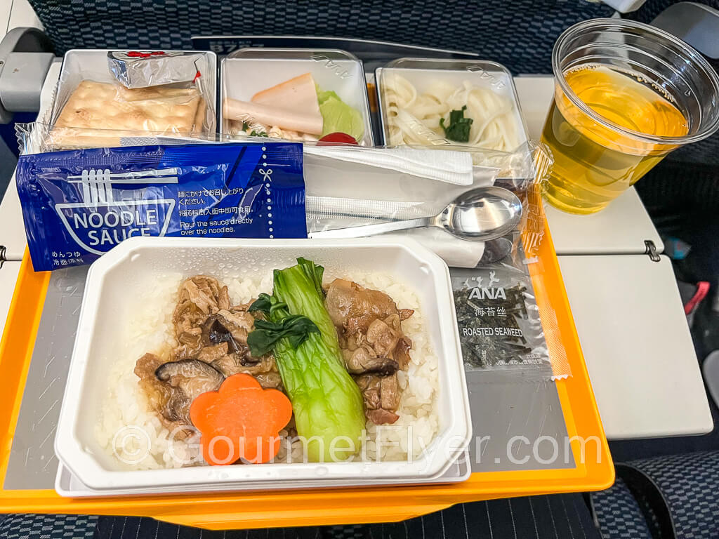 Dinner tray includes the main entree, and three containers of the side dishes line the top of the tray. A roll up of silverware is placed in the middle of the tray.