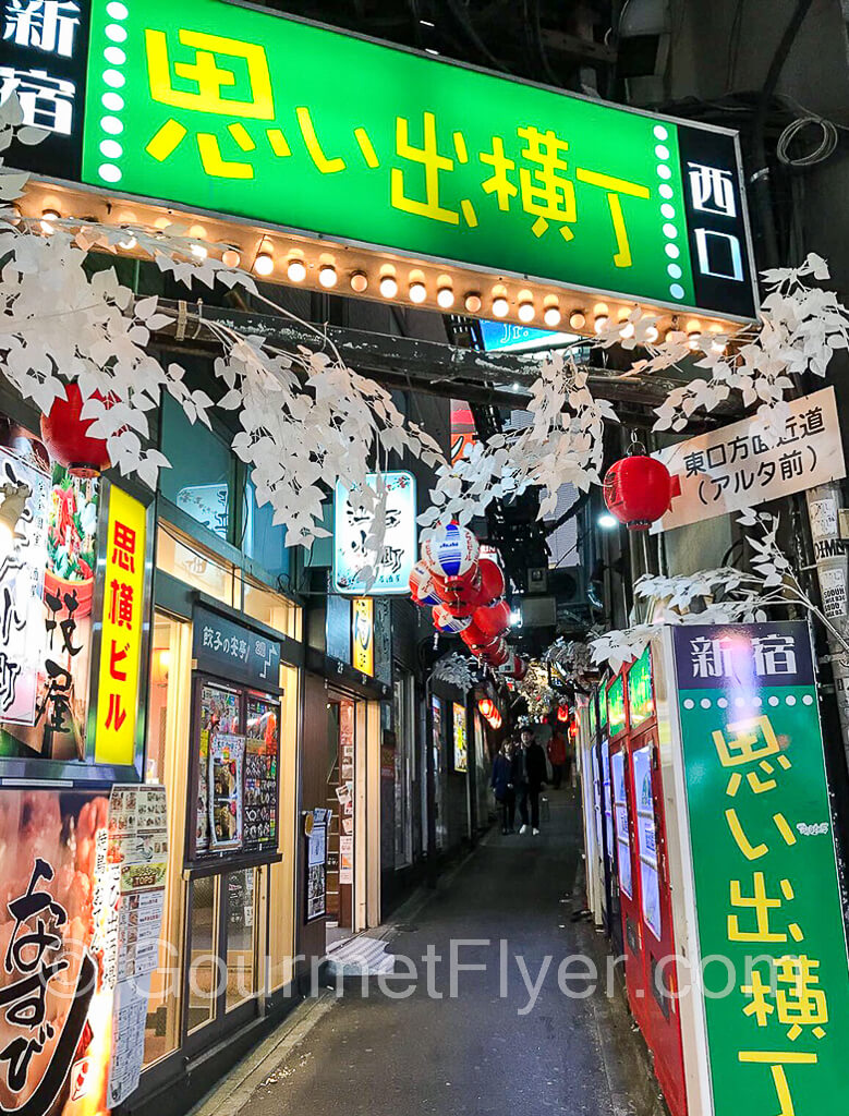 Entrance to a long and dark alley with green and yellow neon signs upfront. A row of red lanterns is hanging above.