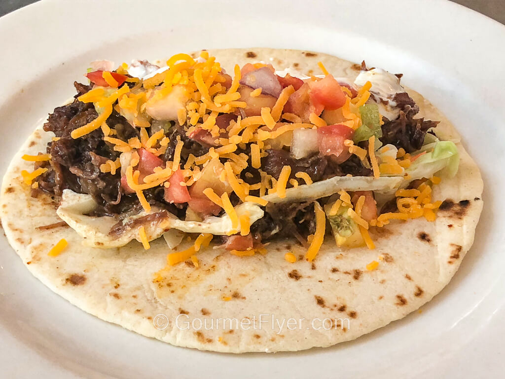 A small flour tortilla on a plate is topped with meat, cheese, and a blend of salsa.