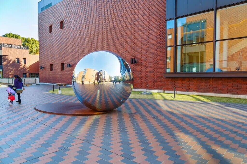 A metallic mirrored spherical sculpture lies in front of a red brick wall with large windows.