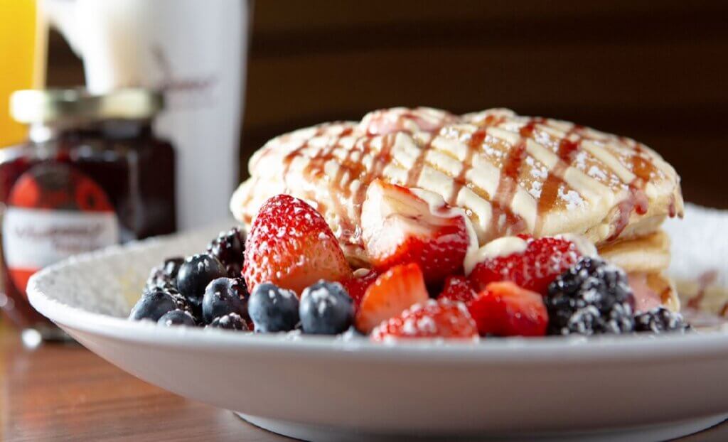 A stack of pancakes sprinkled with powder sugar and topped with syrup is garnished with a medley of berries.