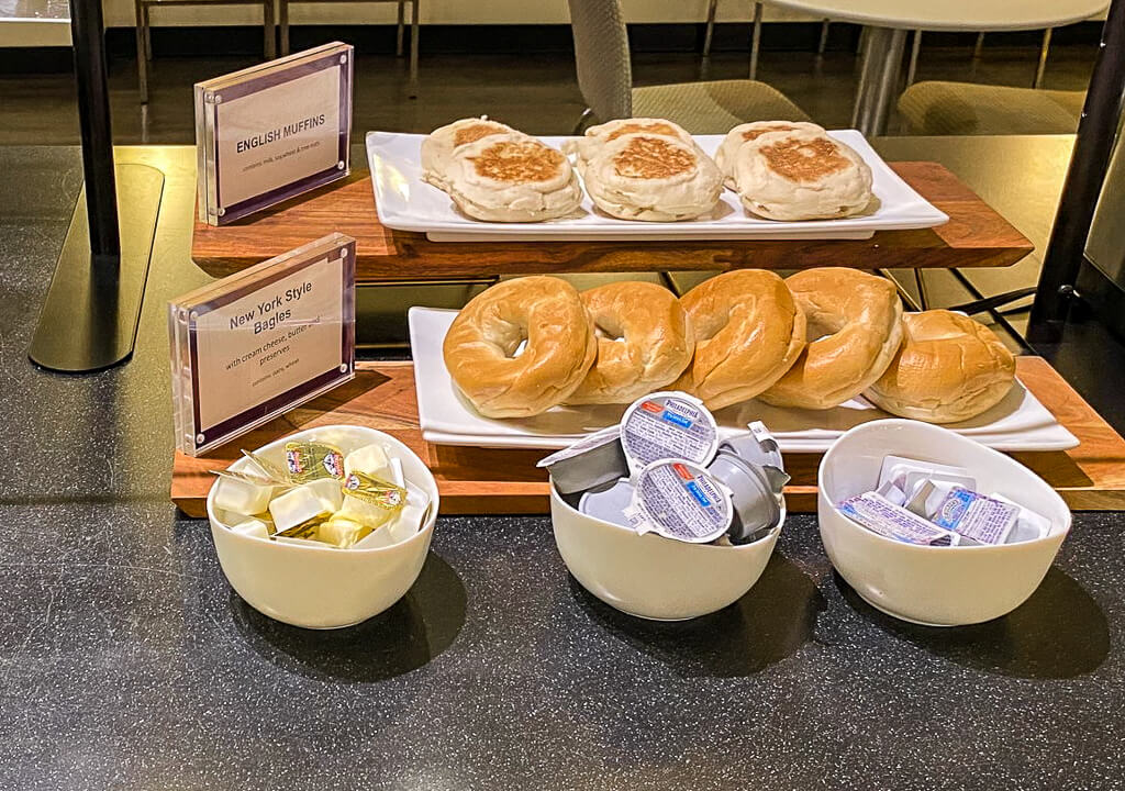 Breakfast items are arranged on the counter with English muffins and New York style bagels. There are bowls containing packaged butter, creamed cheese, and jam.