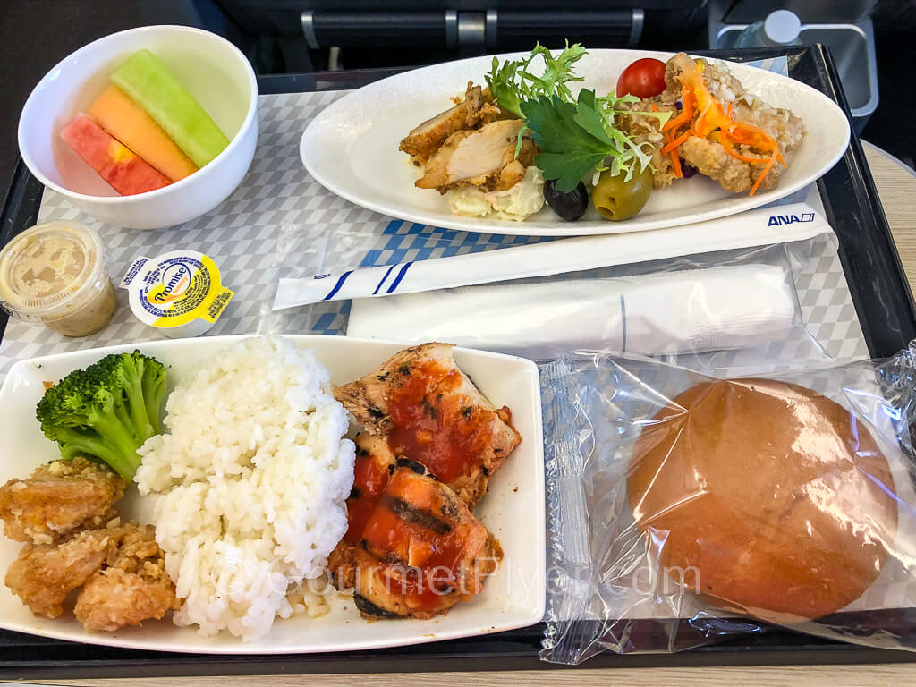 A dinner tray with the appetizer dish on the top of the tray and the main entree on the lower left. A dinner roll and a bowl of sliced fruits complete the service.