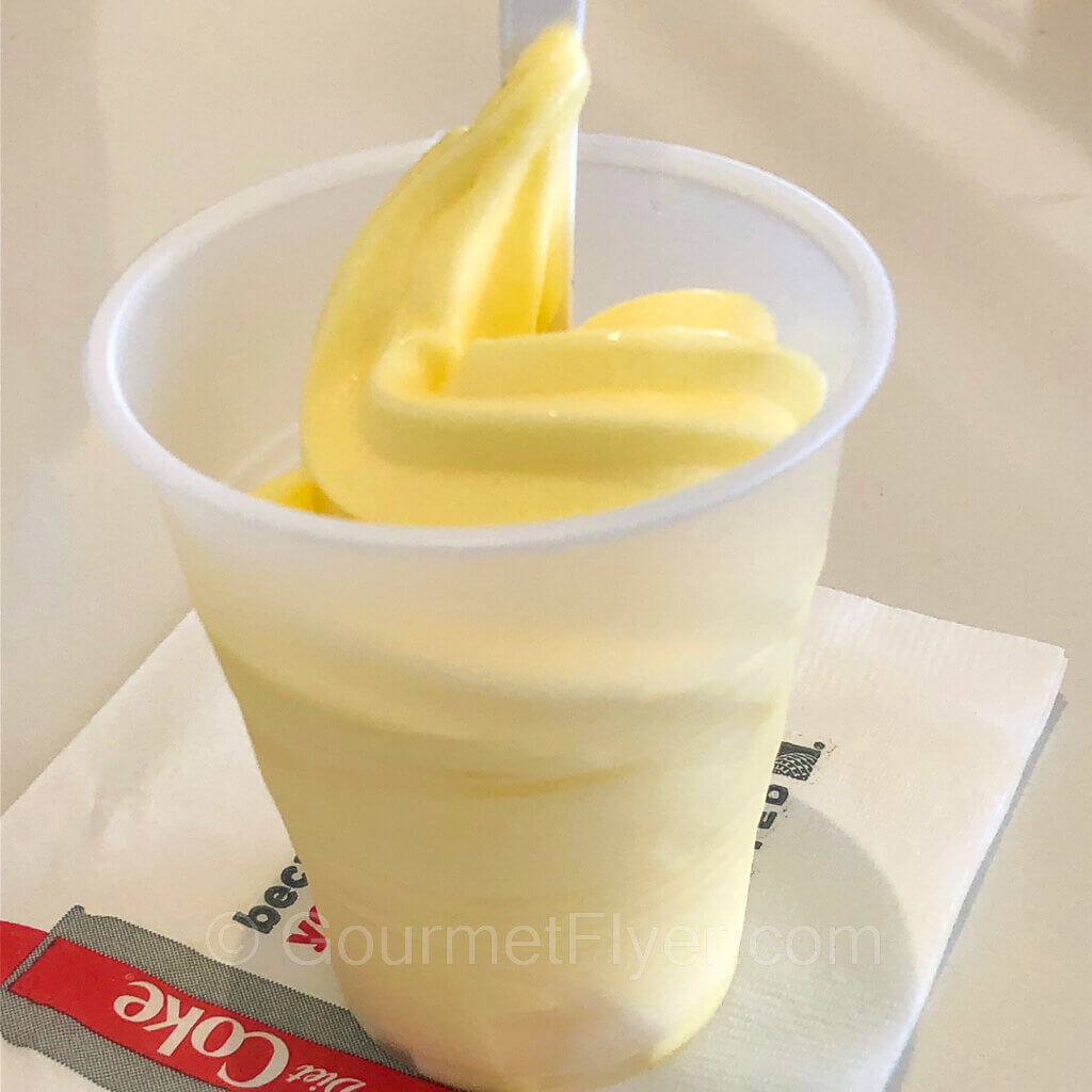Twirling soft pineapple whip served in a tall white plastic cup with a spoon.