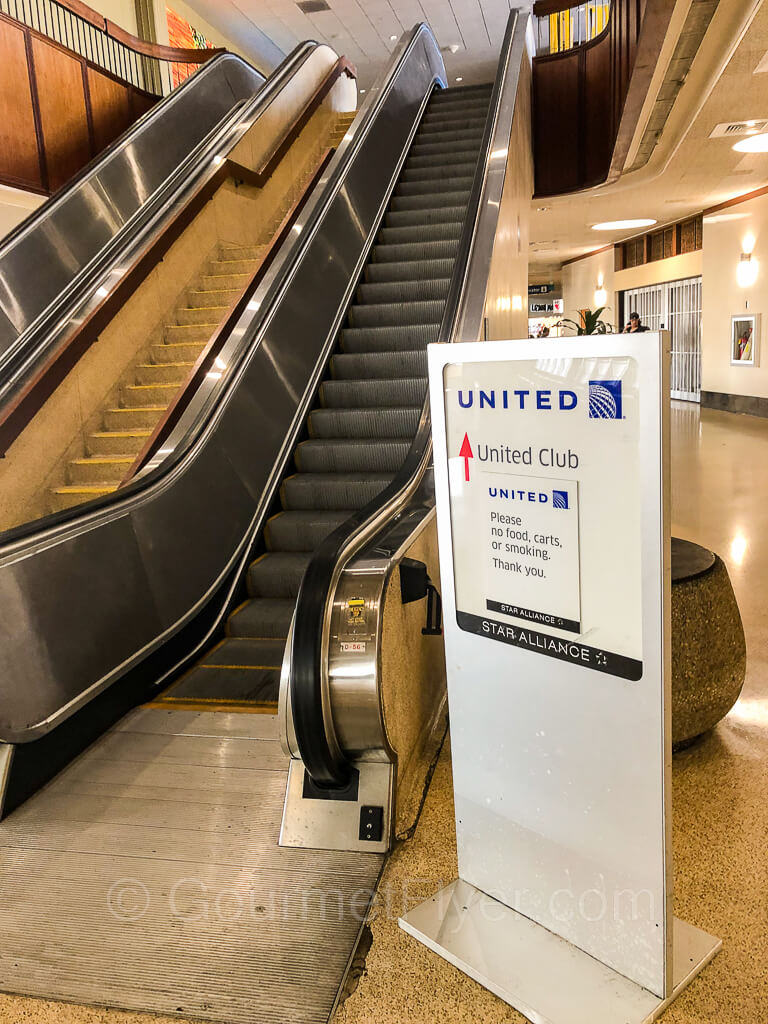 A United Club sign is placed next to a set of two long escalators, with stairs between them.