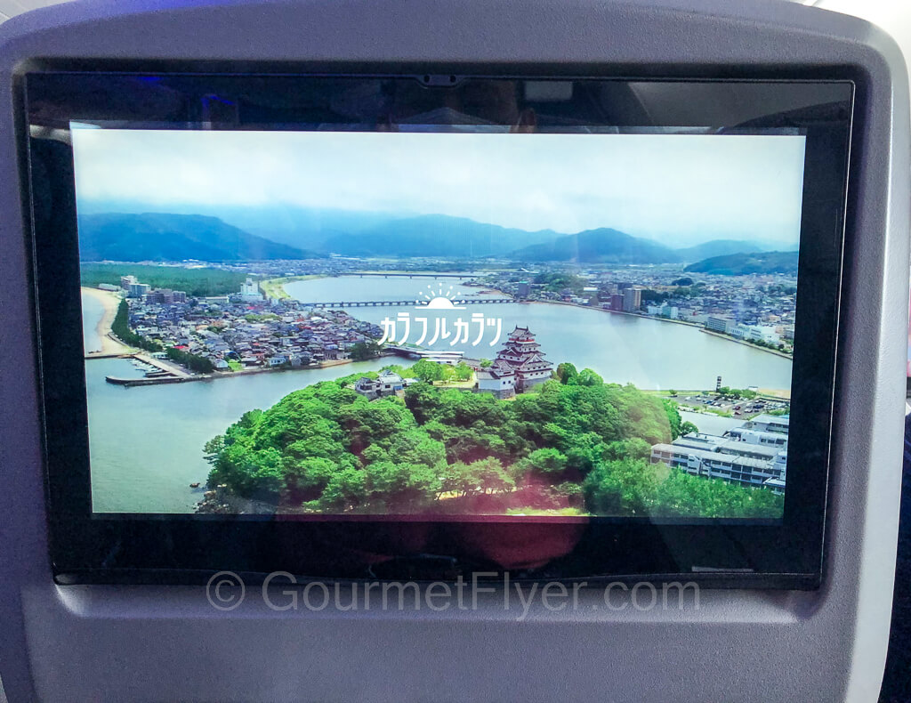 A seatback entertainment screen shows a scenic landscape in a Japanese movie.