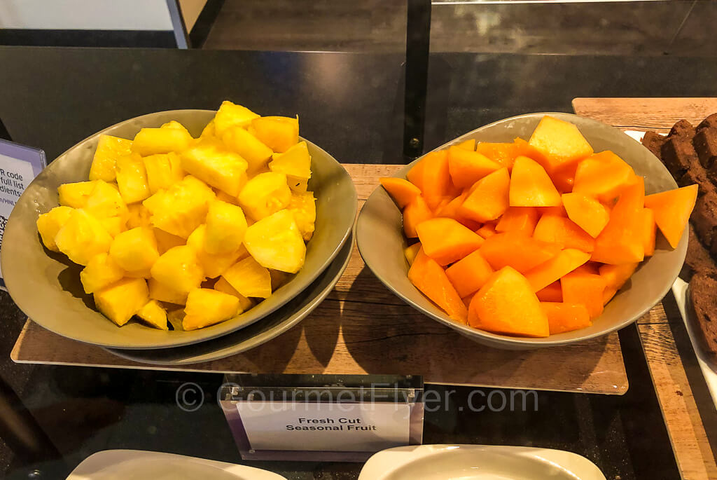 A bowl of cut pineapples and a bowl of cut melons placed side by side on a countertop.
