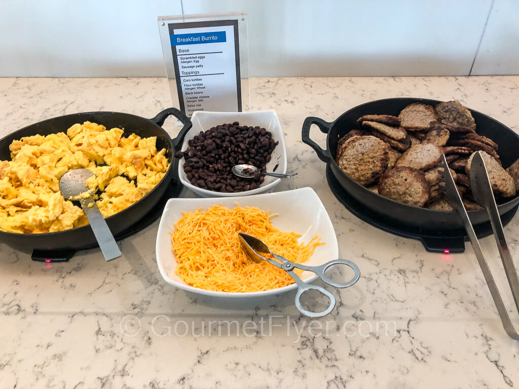 A station with scrambled eggs and sausage patties in iron warmers, along with bowls of shredded cheese and beans.