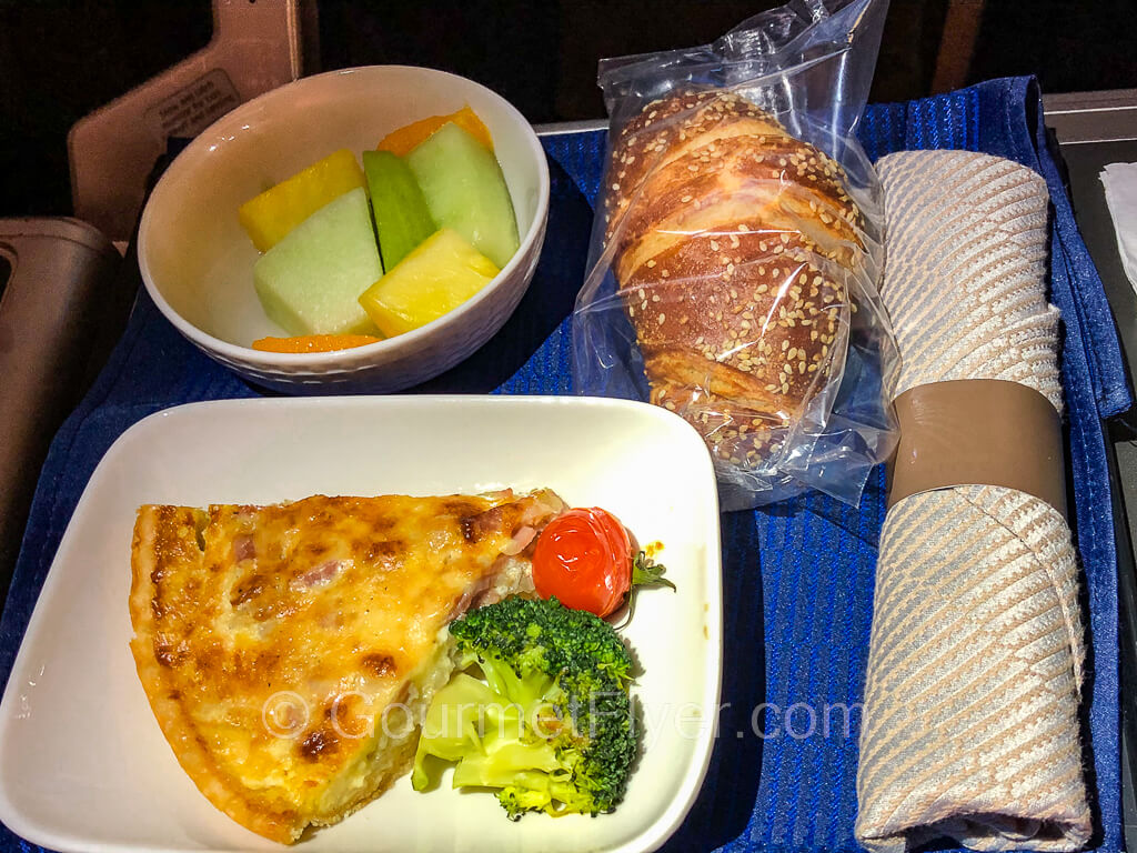 A slice of quiche is garnished with a broccoli floret and a cherry tomato. The plate was served on a tray with blue tablecloth and a silverware rollup.