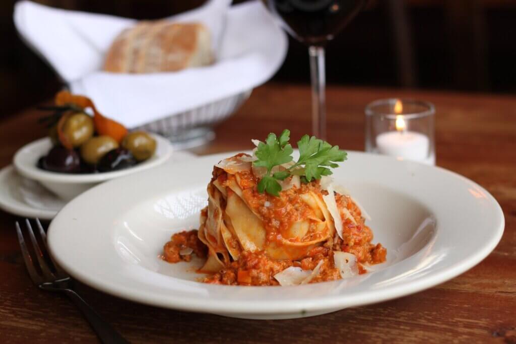 A serving of lasagna served on a white bowl on a wooden dining table with condiments in the backgroud. 