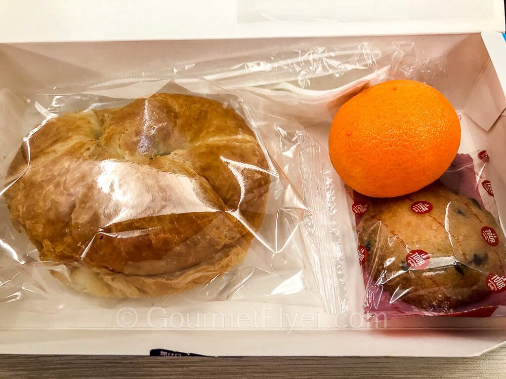 The contents of the snack box are a wrapped croissant sandwich on the left, and a tangerine with a muffin on the right. 