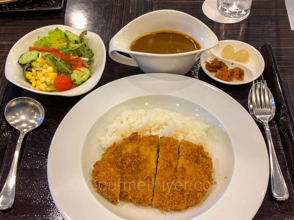 Tonkatsu with rice are served in a plate with curry in a gravy bowl and a side salad in a salad bowl. Silverware accompanies the meal. 
