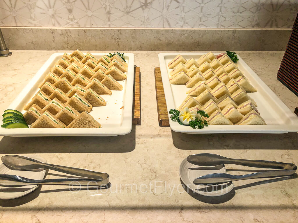 Two long rectangular trays of finger sandwiches are placed on a countertop.