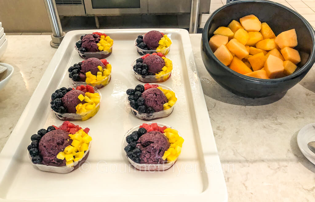 A tray of 8 acai bowls each filled with berries, pineapples, and melons. A bowl of cut melons sits to the right.