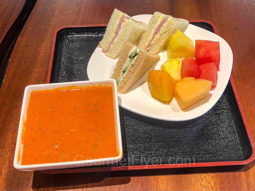 A tray containing a bowl of tomato soup and a plate of finger sandwiches and cut fruits sits on a table.