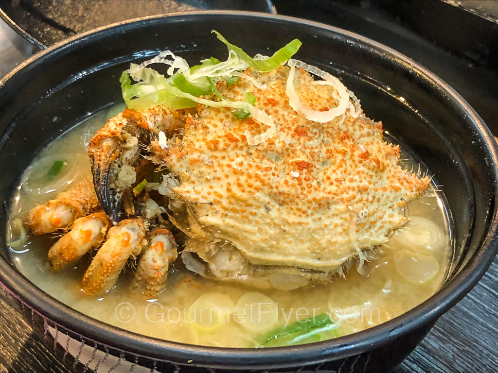 A bowl of miso soup is served with a whole cooked crab and garnished with green onions.