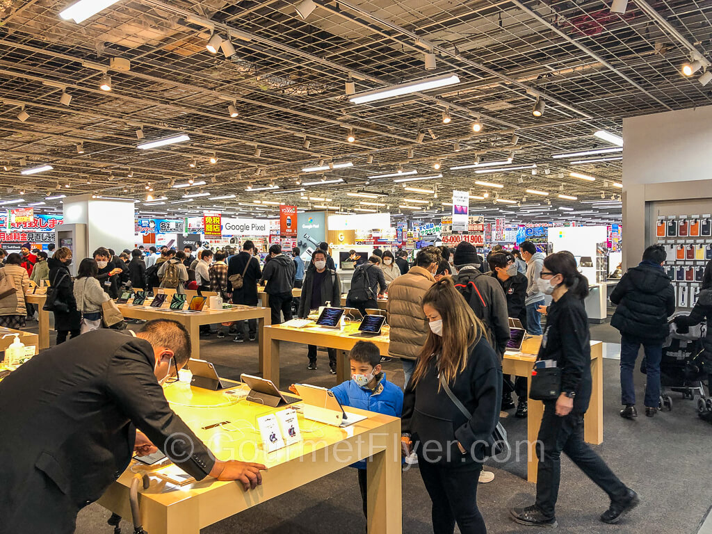 Crowds of shoppers examine tablets on display on multiple large tables in a section of a large electronics store.