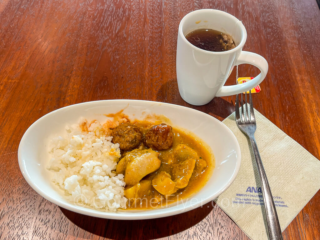 A dish is filled with white rice, curry, and meatballs. A cup of black tea and a fork accompany the dish.