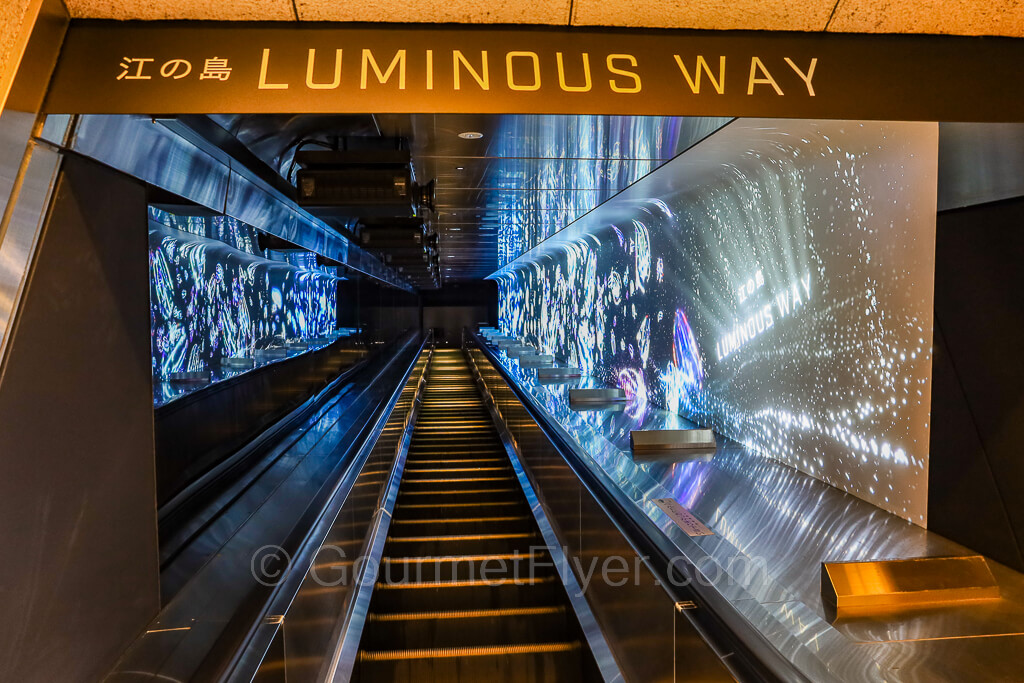 The sign "Luminous Way" hangs above an escalator with illuminations on both walls on the sides.