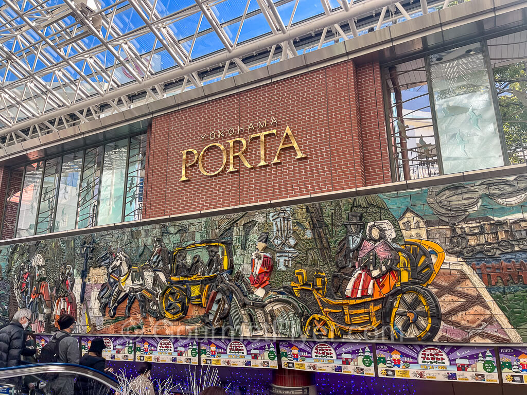 A Yokohama Porta sign sits on a brick background with a large wall of a colorful, and vibrant oil painting under it.