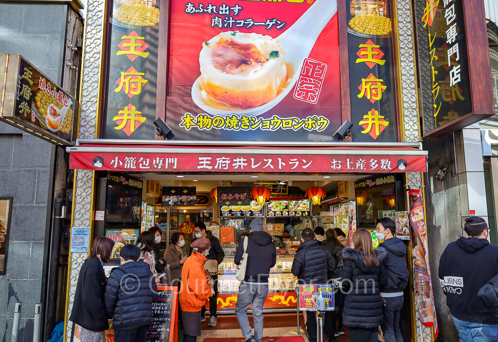 Line forms outside the Chinese restaurant with a large sign of a dumpling with plenty of soup in a spoon.