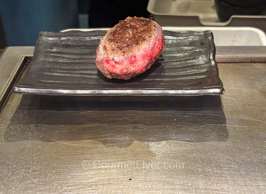 A thick hamburger steak charred on the top side is placed on a metal plate that sits atop a grill.
