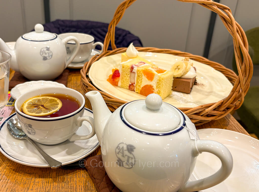 An afternoon tea setting includes cups of tea with lemon, teapots, and a basket of a variety of cakes.