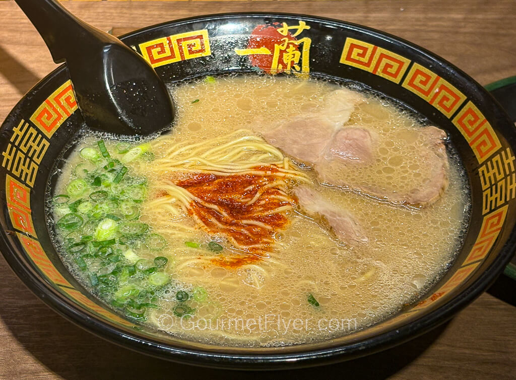 A bowl of ramen is garnished with green onions, topped with sliced meat, and presented with a dot of red chili sauce.