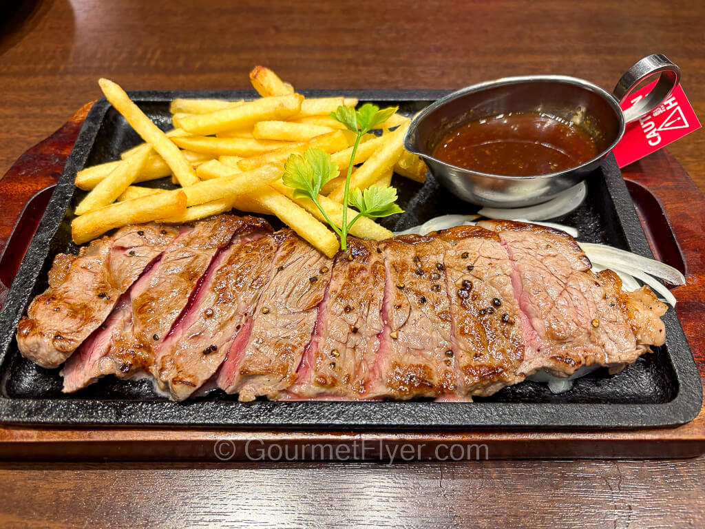 A steak is cut up and served on a metal platter accompanied by fries and a mini gravy boat filled with a wine sauce.