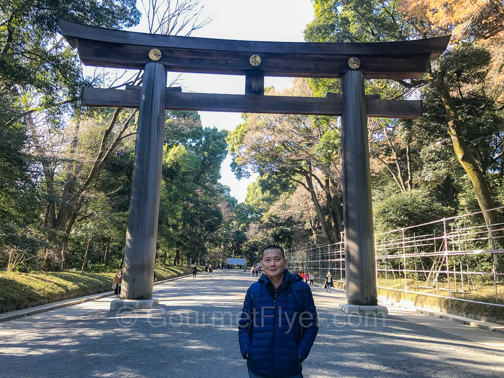 The Gourmet Flyer is posing at the torii gate that marks the entrance to the Meiji Jingu, with many trees in the background.